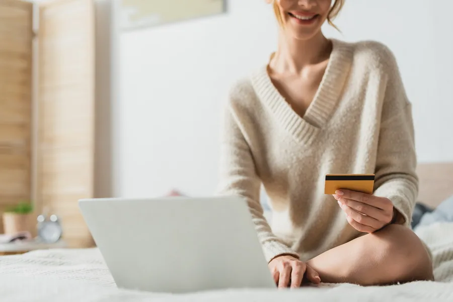 partial view of happy woman holding credit card near laptop while doing online shopping in bedroom